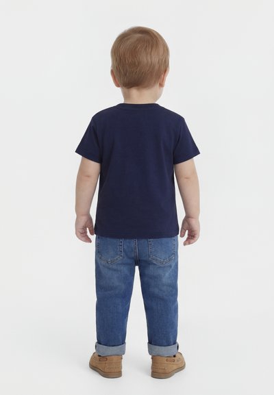 Maloletni otrok stoji obrnjeno stran, oblečen v temno modro majico, zavihane modre kavbojke in rjave čevlje, na belem ozadju.