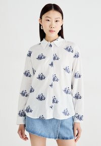 White button-up shirt with navy floral print, long sleeves, pointed collar, and soft texture, paired with a denim mini skirt.