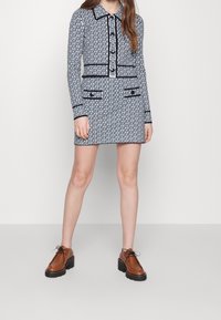 Kobieta ubrana w granatowo-białą sukienkę z długim rękawem w deseń, z czarnymi wykończeniami i guzikami, w połączeniu z brązowymi butami na platformie zawiązywanymi na sznurówki.