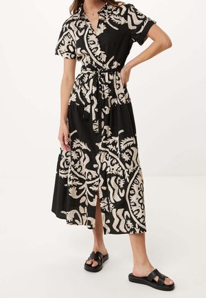 Femme portant une robe midi noire avec des motifs botaniques blancs, manches courtes, boutonnée à l'avant, ceinture à la taille, et sandales noires à enfiler.