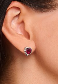 Herzförmiger Rubin-Ohrstecker mit silbernen Akzenten, umgeben von kleinen klaren Steinen, die an einem durchstochenen Ohr befestigt sind.