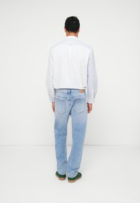 Jeans din denim albastru deschis cu croială dreaptă, asortați cu o cămașă albastru deschis cu butoni, văzuți din spate. Încălțăminte verde.