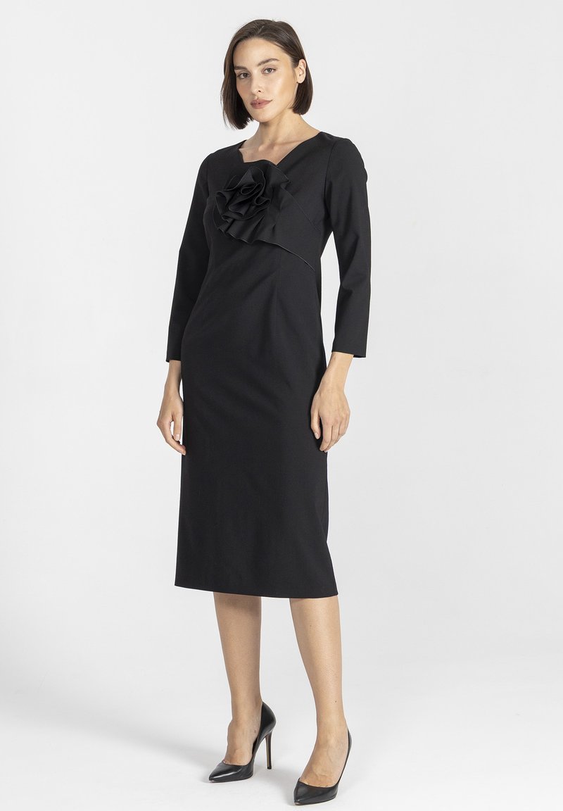 Czarna dopasowana sukienka z dekoltem w serek, rękawami o długości trzech czwartek i dużą falbaną na biuście. Gładki materiał i długość do kolan.