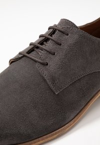Chaussure en daim gris à texture lisse, avec cinq œillets et des lacets marron. La semelle est marron clair avec une forme légèrement arrondie.