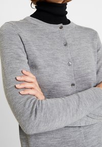 Grå stickad cardigan med rund hals och långa ärmar, prydd med fem svarta knappar fram och en slät textur.