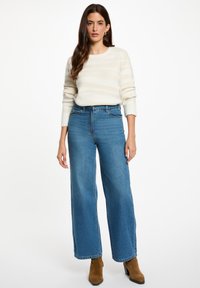 Suéter de rayas crema con cuello redondo y mangas largas, combinado con pantalones vaqueros de pierna ancha de talle alto en azul y botas de tobillo color beige.