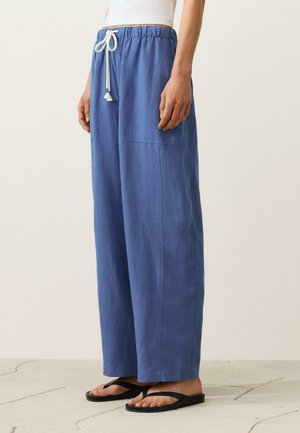 Personne portant un pantalon bleu ample avec un cordon blanc et des tongs noires, debout sur un sol carrelé.