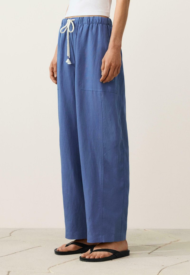 Personne portant un pantalon bleu ample avec un cordon blanc et des tongs noires, debout sur un sol carrelé.