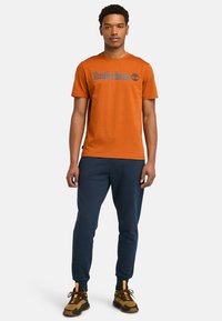 Orange bomulds t-shirt med "Timberland" logo i blå, parret med marineblå sweatpants og sorte/gule sneakers. Enkel crew neck design.