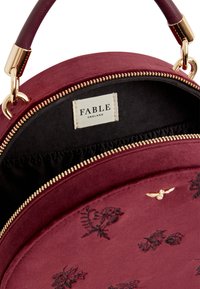 Ronde bordeaux tas met een gladde textuur, gouden rits en zwarte binnenkant. Kenmerkt zich door bloemmotieven in reliëf en een FABLE-label aan de binnenzijde.