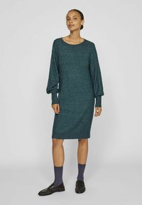 Abito lavorato a maglia in verde acqua, di lunghezza media, con scollo rotondo e maniche a palloncino. Il tessuto testurizzato e i polsini a coste aggiungono un tocco di dettaglio. Indossato con calze scure e scarpe.