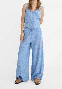 Lichtblauwe linnen vest met vijf knopen, gecombineerd met een bijpassende broek met wijde pijpen en een trekkoord in de taille. Casual, ademend materiaal.