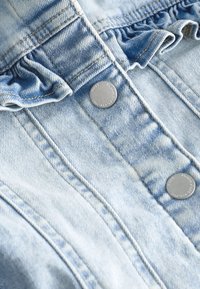 Svetlomodrá rifľovina s vertikálnym prešitím, striebornými patentkami a volánovým detailom pri hornom okraji.