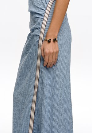 Mano de mujer que lleva una pulsera dorada con flores, apoyada junto a una falda azul texturizada con una raya vertical beige y marrón.