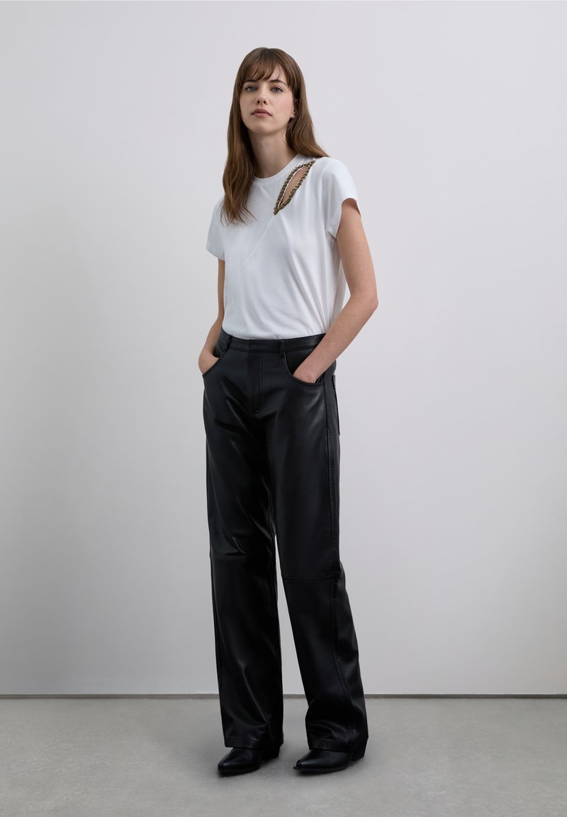 White short-sleeve t-shirt with a diagonal design and chain detail, paired with wide-leg black leather pants, worn with black heeled boots.