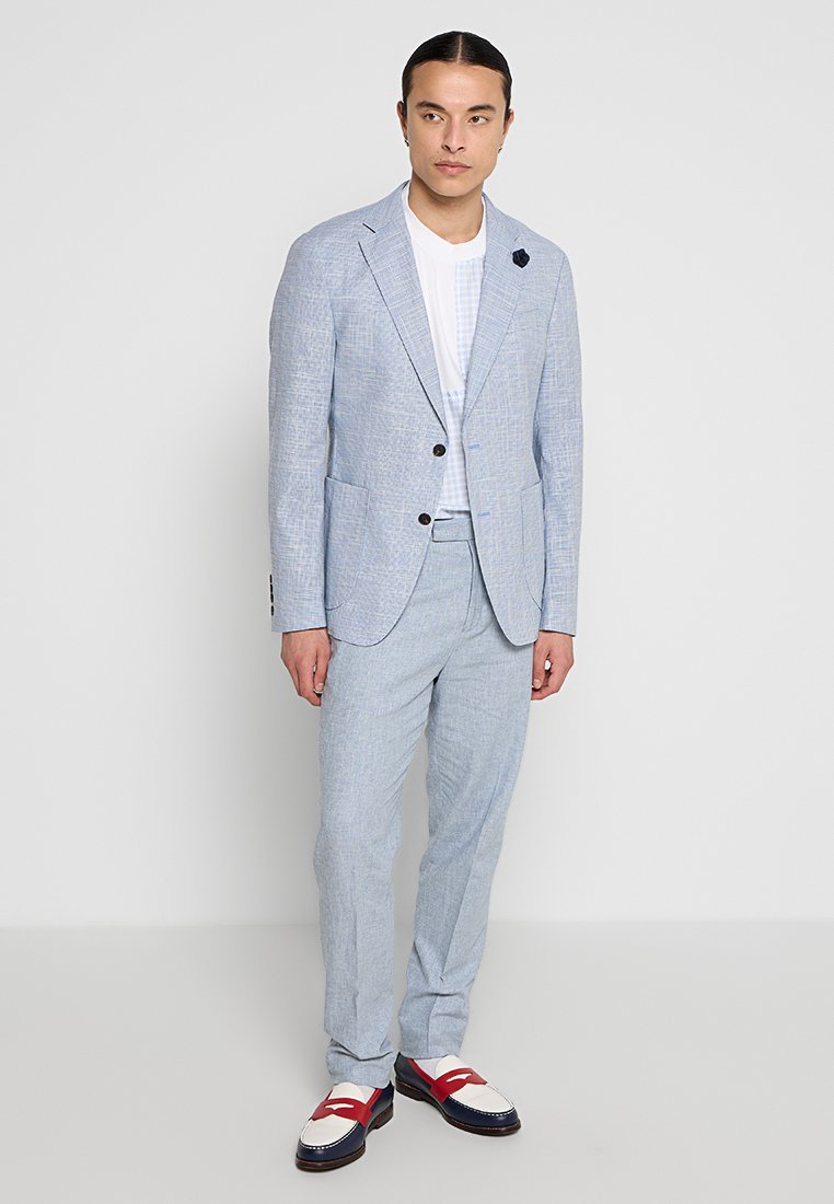 Completo su misura azzurro chiaro con un motivo testurizzato, abbinato a una t-shirt bianca e scarpe a doppia fibbia con dettagli rossi.