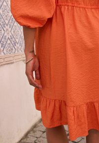 Orange, rynkigt klänning av tyg med puffärmar och en volangkant. Hand med ett guldarmband som vilar på klänningen. Texturerad design.