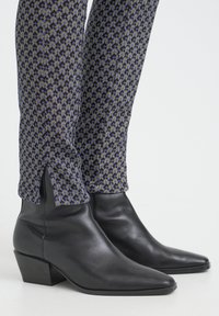 Schwarze Leder-Stiefeletten mit spitzer Zehenpartie und niedrigem Blockabsatz, kombiniert mit gemusterten, eng anliegenden blauen und gelben Hosen mit seitlichen Schlitzen.