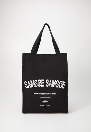 Sac cabas noir avec de larges poignées présentant un texte blanc déformé "SAMSØE SAMSØE" et un texte plus petit "When birds sing we whisper" ainsi qu’une adresse à Copenhague.