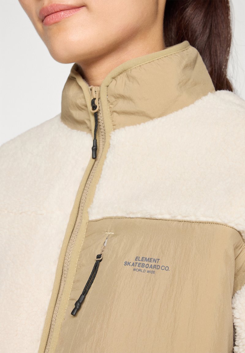 Giacca beige foderata in pile con colletto alto e chiusura con zip. Presenta una stampa del logo sul petto e trame contrastanti di tessuti lisci e soffici.