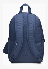Sac à dos bleu marine avec des bretelles rembourrées, arborant un logo "CONVERSE" en relief et une poche latérale pour le rangement. Texture de tissu durable.