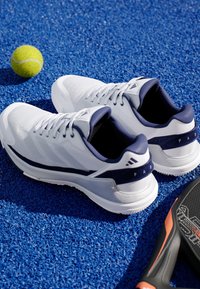 Bílé sportovní boty s námořnickými akcenty, síťovaným vrškem a gumovou podrážkou. Umístěné na modrém texturovaném povrchu vedle zeleného tenisového míčku a rakety.