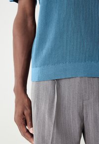 Kortärmad ribbad stickad tröja i ljusblått, kombinerad med ljusgråa randiga byxor. Närbild på en hand som vilar på midjan.