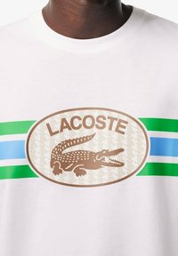 Camiseta de algodón blanca con un gran gráfico ovalado. Texto "LACOSTE" sobre un logotipo de cocodrilo marrón, con acentos de rayas verdes y azules.