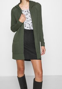 Olivově zelený pletený kardigan s kapucí, s žebrovanými manžetami a dvěma předními kapsami, nošený přes vzorované šedé tričko a černou sukni.