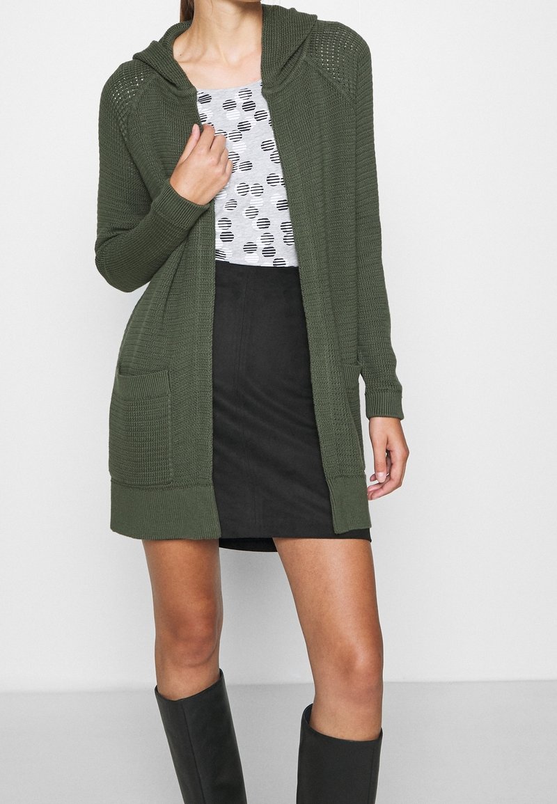 Olivově zelený pletený kardigan s kapucí, s žebrovanými manžetami a dvěma předními kapsami, nošený přes vzorované šedé tričko a černou sukni.