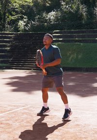Tenis igralec v temno zeleni majici s kratkimi rokavi in sivih hlačah, ki drži rdeč tenis racket na peščeni igri, obdani z zelenjem.