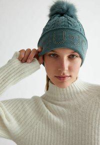 Berretto lavorato a maglia color turchese con pompon e borchie decorative che formano la scritta "LIU•JO." La modella indossa un maglione a collo alto a coste color crema.
