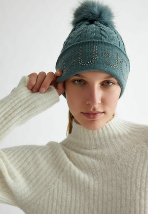 Berretto lavorato a maglia color turchese con pompon e borchie decorative che formano la scritta "LIU•JO." La modella indossa un maglione a collo alto a coste color crema.