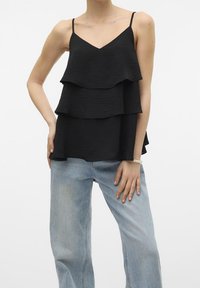 Camisola nera a strati con tre strati arricciati, spalline sottili e scollatura a V, abbinata a jeans a gamba larga azzurri.