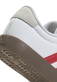 Zapatilla blanca con rayas rojas, detalles en gamuza gris en el talón y una suela de goma marrón texturada que muestra un patrón de tacos.