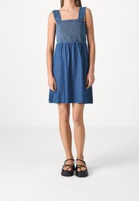 Vestido de mezclilla con un corpiño fruncido y escote cuadrado, que presenta tirantes anchos y una falda de corte suelto. El color es un azul medio.