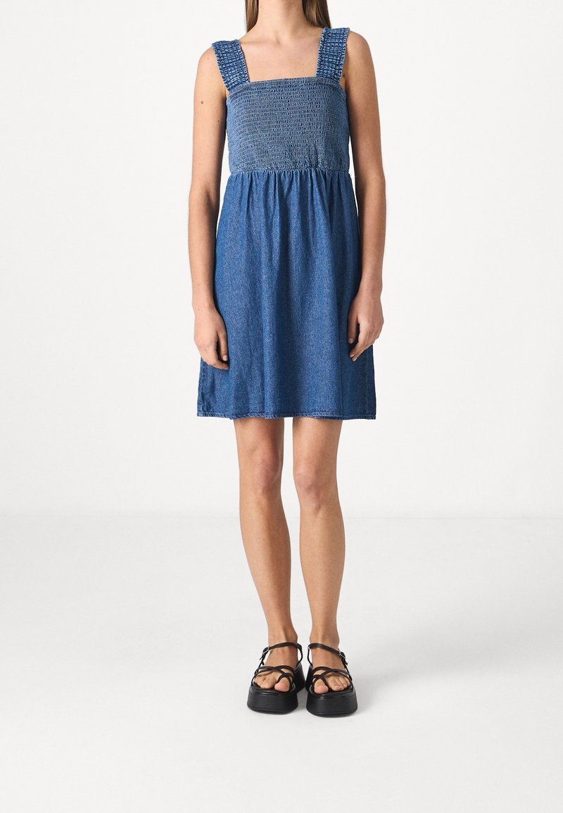 Vestido de mezclilla con un corpiño fruncido y escote cuadrado, que presenta tirantes anchos y una falda de corte suelto. El color es un azul medio.