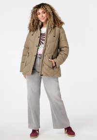 Bruine, gestikte pufferjack met een hoge kraag, ritszakken en een aansluitend ontwerp, gedragen over een graphic tee en in combinatie met grijze wijde jeans.