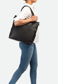 Borsa tote nera in pelle con due manici, dotata di una tasca frontale con zip e finitura testurizzata; indossata da una persona in jeans attillati e camicia bianca.