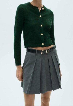 Femme portant un cardigan à manches longues vert foncé avec des boutons dorés et une mini-jupe plissée grise avec une ceinture noire à boucle dorée.