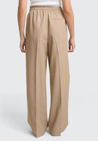 Pantaloni beige a gamba larga con cintura elastica e tasca posteriore indossati da una persona in piedi contro uno sfondo neutro.
