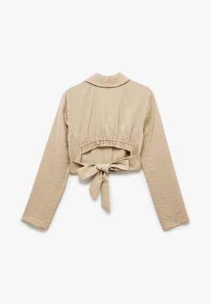 Veste beige courte avec manches longues, col, ceinture élastique et un détail noué au dos. Texture de tissu lisse et léger.