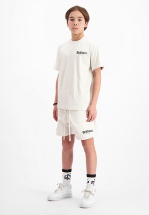 Lichtbeige t-shirt en shorts set met een subtiele textuur, voorzien van een logo op de borst en dij. Draag met witte sneakers en gestreepte sokken.