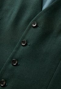 Tamsiai žalia vilnos švarkas su keturiais juodais mygtukais, pritaikytomis atvartais ir lygiu paviršiumi su subtiliomis siuvimo detalėmis.