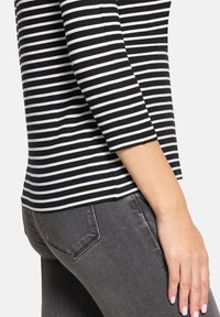 Schwarz-weiß gestreiftes Oberteil mit Dreiviertelärmeln; kombiniert mit grauen Jeans. Der Stoff scheint weich mit einer glatten Textur und tailliertem Design zu sein.