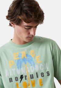 Camiseta de algodón verde claro con texto gráfico multicolor y un diseño de palmera, con acentos en azul, amarillo, gris y negro.