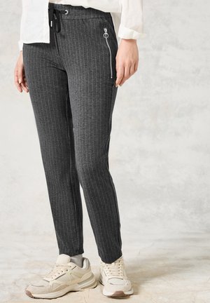 Femme portant un pantalon à fines rayures gris foncé avec une poche zippée et des baskets beiges, debout contre un mur gris clair.