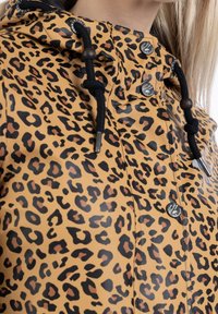 Lepardmönstrad regnjacka i beige och svart, tillverkad av vattentåligt material, med huva, dragsko och metalltryckknappar.