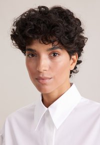 Weißes Hemd mit glatter Textur. Das Model hat kurzes, lockiges schwarzes Haar. Natürliche Schminke, neutraler Hintergrund. Nahaufnahme.