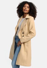 Beige wollen jas met een dubbelgeknoopte sluiting, een reverskraag en zijzakken, gecombineerd met lichtblauwe jeans en een wit topje.